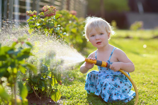 Cute toddler girl watering flowers from hose in summer garden