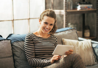 Obraz premium Smiling young woman sitting on sofa and using tablet pc
