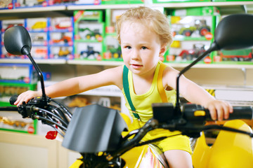 Little cute boy in yellow sits on toy motorcycle in store