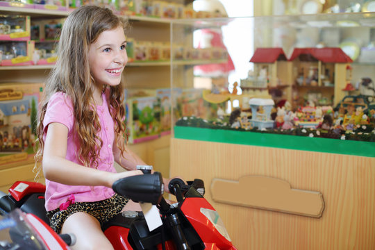 Little cute girl in sits on red toy motorcycle in store with toy