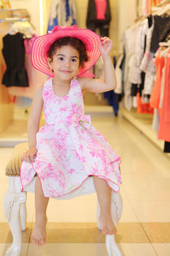 Little barefoot girl in hat sits in children store with clothes