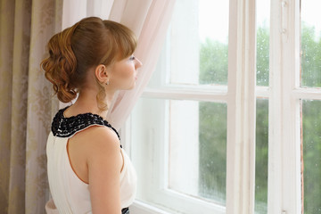 Beautiful sad girl in white dress looks in window in light room.