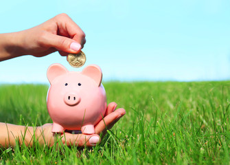 Piggy Bank and Coin in Female Hands over Green Grass
