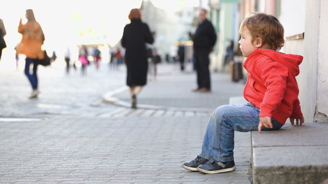 Little boy sitting alone in the street, people passing by