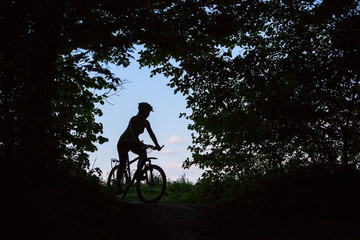Silhouette of woman that stands with bike at the forest edge