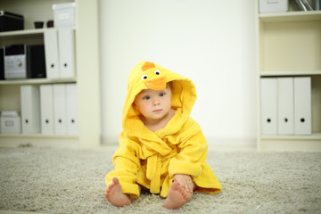 Little cute baby in yellow robe sits on white carpet and looks
