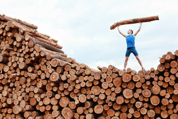 man on top of large pile of logs, lifting heavy log