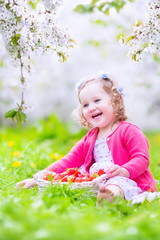 Pretty toddler girl eating strawberry in blooming garden