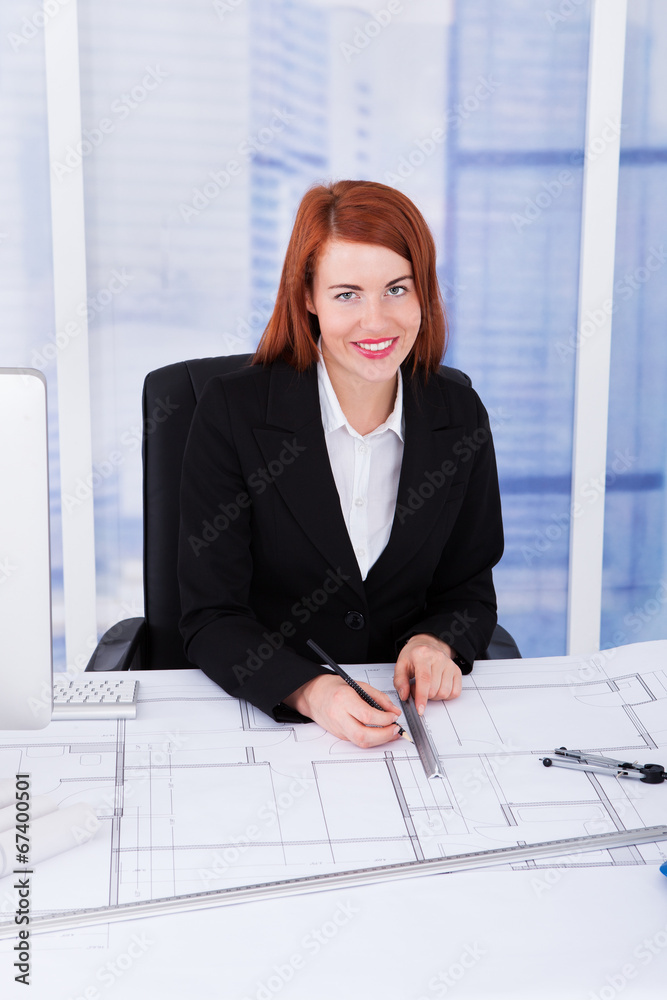 Poster businesswoman using computer with blueprints at desk