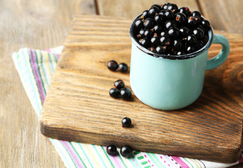 Ripe blackcurrants in mug on board, on wooden background.