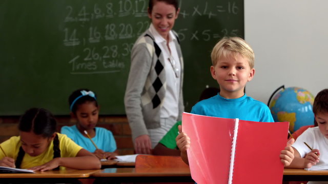 Little boy holding red notepad smiling at camera in classroom