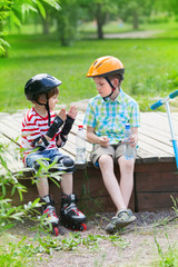 two boys with rollers and kick scooter sit in the park