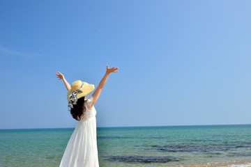 Fototapeta na wymiar 麦わら帽子と白いドレスを着てビーチに立っているアジア人女性の後ろ姿