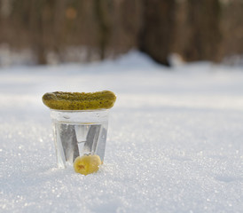 Рюмка водки и закуска - огурец с оливкой на снегу