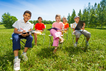 Kids hold sketch-boards and sit outside on chairs