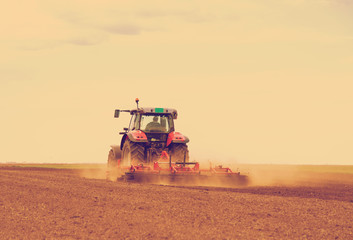 Naklejka premium Red tractor driven by farmer cultivating land at spring