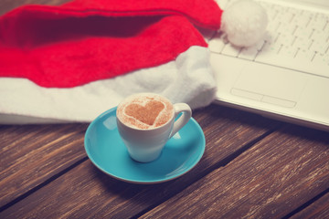 Cup of coffee with heart shape, laptop and christmas hat.