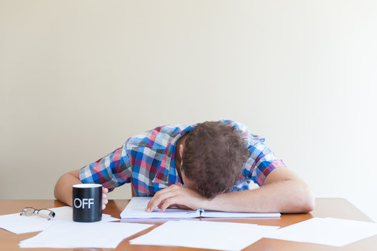 young adult asleep over books holding a cup.