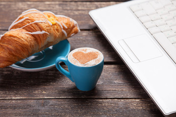 Croissant and cup of coffee with laptop on wooden table.