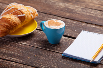 Croissant and cup of coffee with notebook on wooden table.