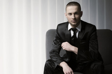 elegant young fashion man in tuxedo on a sofa, blinds background