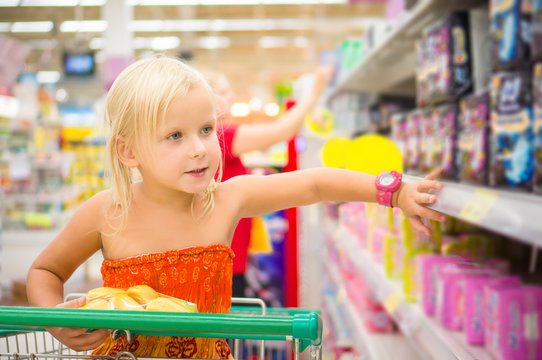 Adorable girl in shopping cart looks at goods on shelves in supe