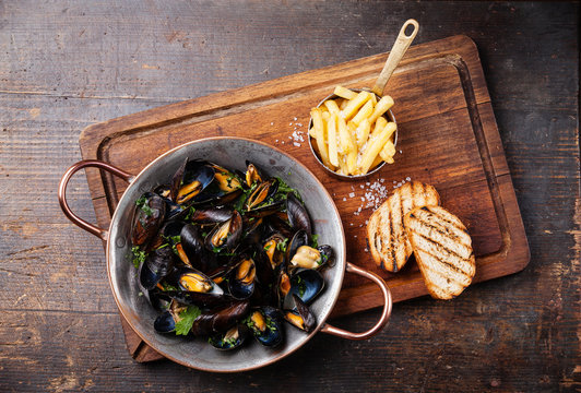 Mussels in copper cooking dish and french fries on dark wooden b