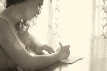 young woman sitting and writing letter near bright window light.