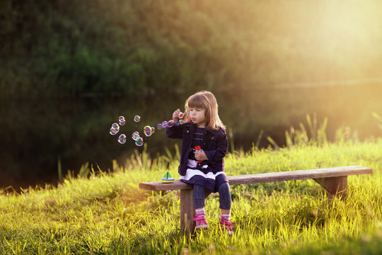 little girl sitting on a wooden bench blows bubbles in the rays