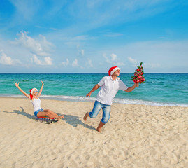 Young couple running at sea beach in santa hats with sled