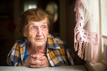 Old woman sitting at a table near the window in his house.