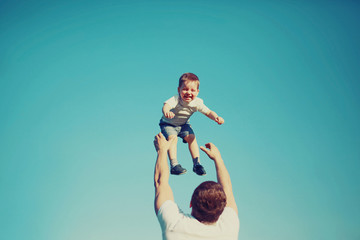 Happy father and son having fun, dad throwing child in the air outdoors, blue sky background,...