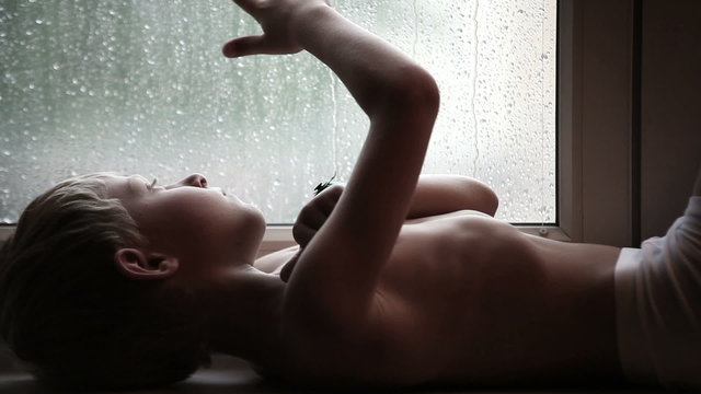 Sad boy on the windowsill watches the raindrops on the glass