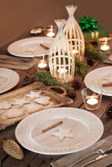 Christmas table setting. Elegant white plates with cookies, natural pine tree branch, pinecones, candles and gifts. Rural or rustic style decorations.