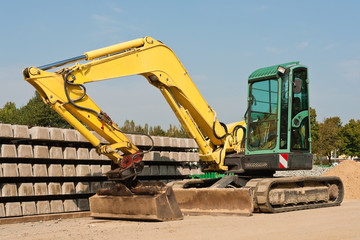 Ein Kompaktbagger steht vor Betonschwellen unter blauem Himmel