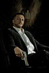 Elegant man sitting in a chair holding  gun over dark background - obrazy, fototapety, plakaty
