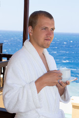 happy man in bathrobe with cup of coffee standing on balcony