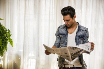 Handsome young man at home reading newspaper, standing