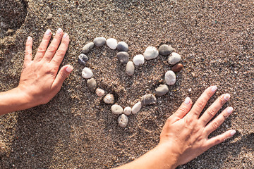 Woman's hands making heart of marine shingles on the sand.