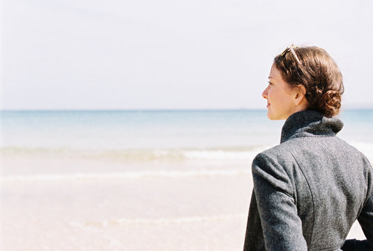 A woman in a grey jacket looking out over a beach and the sea.