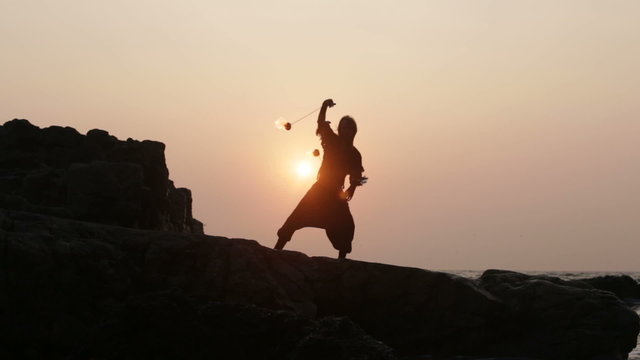 man turns fire poi standing on a cliff near the ocean at sunset