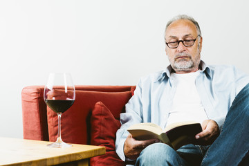 Rentner sitzt auf Sofa, liest Buch, Rotweinglas