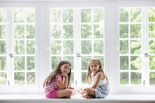 Two girls with digital tablet sitting by large window