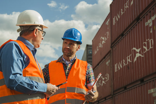 Two men with safety helmets and reflective vests talking at container port