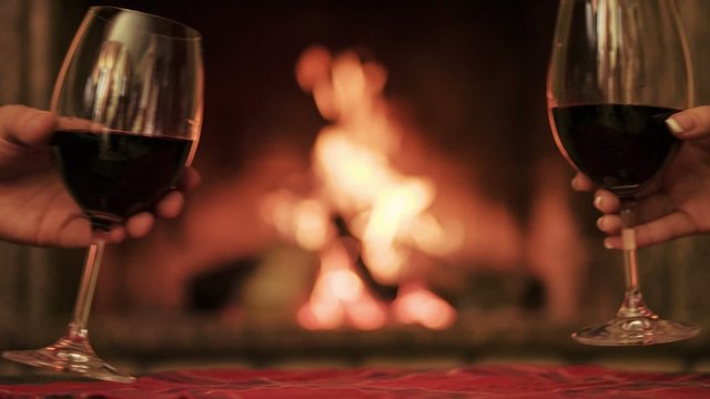 Couple toasting with red wine by the cozy fireplace. Close up.