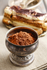 Silver goblet of cocoa powder. Tiramisu cake in the background