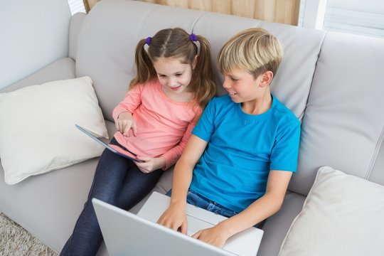 Happy siblings using laptop and tablet pc on sofa