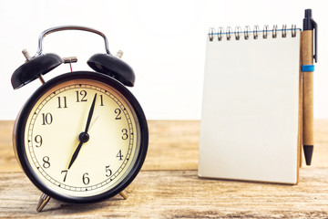 Black old clock and blank paper with pen, on wooden table.