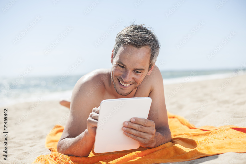 Wall mural handsome man on the seaside lying on a beach towel to use a digi