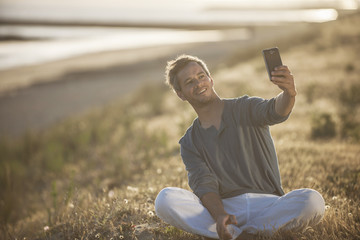 handsome man sitting on the beach at sunset and taking a selfie